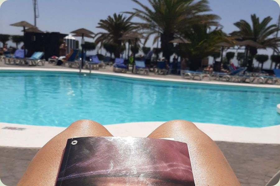 Een persoon ontspant bij een zwembad met palmbomen en leest een boek dat op de buik rust, onder de Spaanse zon in Alicante.