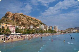 Een breed, zanderig strand, vol met badgasten, grenst aan turkooisblauw water. Achter het strand staan rijen hoge appartementen en palmbomen, die zich uitstrekken tot aan de basis van een grote, steile, rotsachtige heuvel. De heuvel wordt bekroond door een historisch kasteel en vestingmuren. De lucht is deels bewolkt.