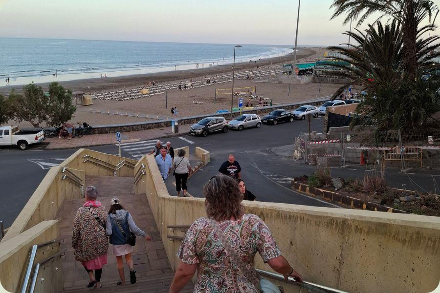 Verhoogd uitzicht in de avond over een breed zandstrand in Alicante met rijen ligbedden en parasols, een beachbar en geparkeerde auto's langs de kustweg. Mensen lopen op de trap en het strand.