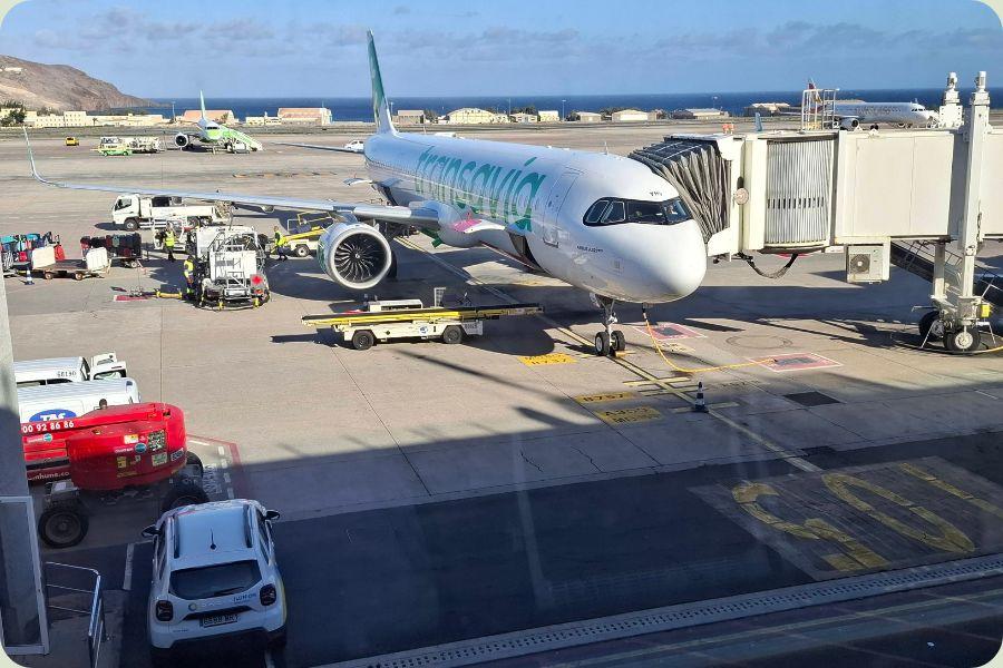 Een Transavia vliegtuig aan de gate op een zonnig vliegveld met zicht op de zee en bergen op de achtergrond, klaar voor vertrek naar Alicante.