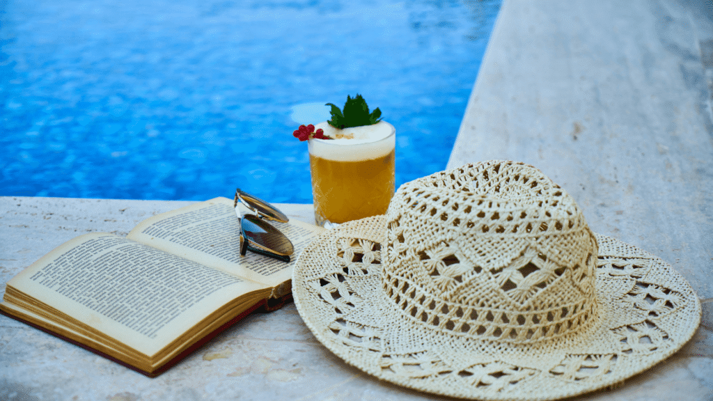Cocktailglas met hoed, zonnebril en boek op een ligbed bij het zwembad van een luxe adults only resort aan zee.