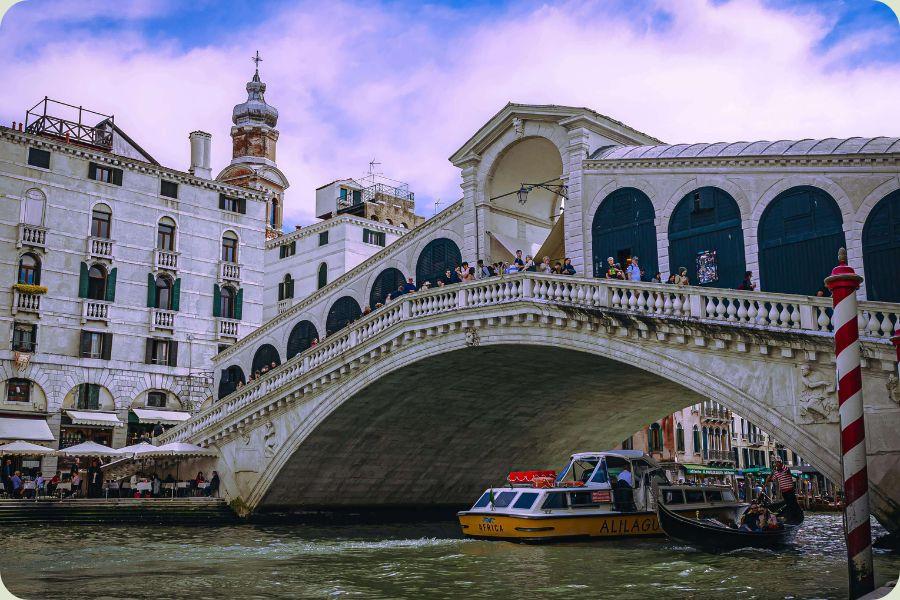 Een levendig zijaanzicht van de stenen Rialtobrug in Venetië met toeristen op de boog, een vaporettoboot die eronderdoor vaart en historische gevels aan de waterkant onder een bewolkte paarsblauwe lucht.